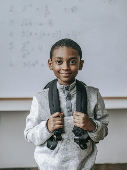 Cheerful African American boy in casual outfit with backpack standing against whiteboard in classroom and looking at camera